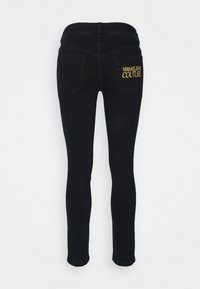 Black skinny jeans shown from the back with two pockets, one featuring gold "Versace Jeans Couture" embroidery on a plain background.