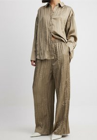 Jasnobrązowa, plisowana koszula z guzikami o luźnym kroju, zestawiona z dopasowanymi spodniami w szerokim kroju. Materiał wydaje się satynowy, o gładkiej fakturze.