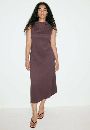 Femme aux longs cheveux bouclés portant une robe midi sans manches violet foncé et des sandales noires, debout devant un fond blanc uni.