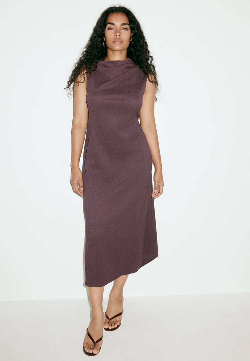 Femme aux longs cheveux bouclés portant une robe midi sans manches violet foncé et des sandales noires, debout devant un fond blanc uni.