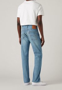 Vaqueros de mezclilla azul claro con ajuste relajado, bolsillos traseros visibles y un parche de cuero marrón en la cintura. Combinados con una camiseta blanca.