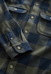 Flannel shirt in green and navy blue check pattern, featuring four buttons, two chest pockets, and textured fabric. Smooth cuffs included.