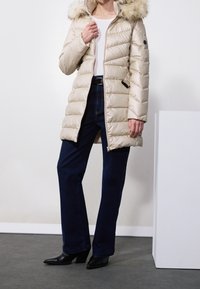Žena v béžovém péřovém zimním kabátu s kapucí lemovanou kožešinou, tmavě modrých džínech, černých podpatcích a bílém topu uvnitř.