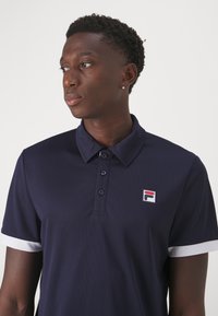 Marine polo skjorte med teksturert stoff, to-knapps placket og hvite detaljer ved ermene. Har en liten merkevarelogo på brystet.