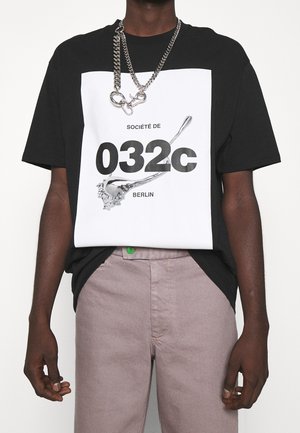 T-shirt graphique noir et blanc avec le texte "Société de 032c BERLIN" et un motif éclaboussure argentée, porté avec un pantalon rose clair. Collier chaîne argentée.