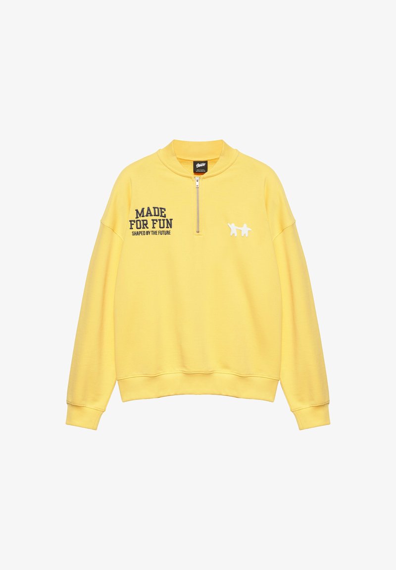 Sweat-shirt jaune à manches longues avec une fermeture éclair à l'avant, texte noir "MADE FOR FUN SHAPED BY THE FUTURE" sur la poitrine gauche et deux petites figures blanches sur la poitrine droite.