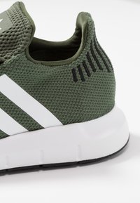 Grön sportskor med en texturerad mesh-övre, vita ränder och en lätt grå sula med svarta accenter och detaljer.