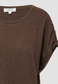 Bruine korte mouwen shirt gemaakt van zacht, textuurstof. Heeft een ronde halslijn en geplooide details op de mouwen. Merkenlabel zichtbaar.