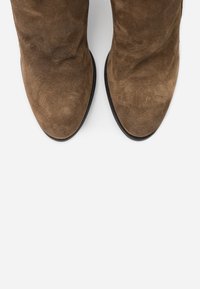 Stivali in suede marrone con punte arrotondate e suole nere su uno sfondo bianco, visti dall'alto.