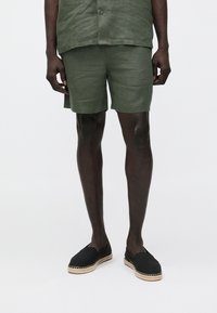 Calções verdes de linho com um corte descontraído, apresentando uma superfície texturizada. Combinados com espadrilhas pretas com detalhes em juta ao longo da sola.