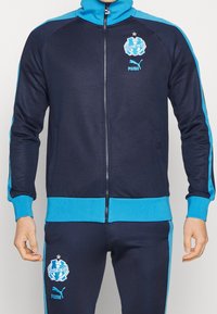 Marinblå zip-up träningsjacka med turkosa detaljer och logotyp. Har ribbade ärmslut och nederkant, och är tillverkad av ett texturerat tyg.