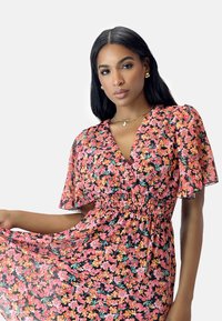 Blumenkleid mit kurzen Puffärmeln, ausgestattet mit einer gerafften Taille. Rosa Hintergrund mit orangefarbenen und grünen Blumenmustern. Leichter, transparenter Stoff.
