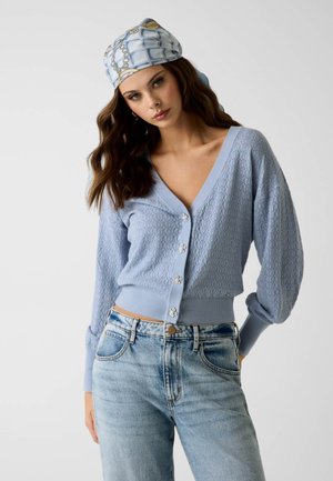 Femme portant un cardigan texturé bleu clair avec des boutons floraux, un jean bleu et un foulard bleu clair à motifs, posant devant un fond uni.