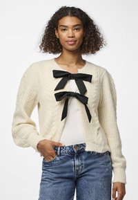 Ελεφαντοστού πλεκτή κάρντιγκαν με υφή από πλέξη καλωδίου, που διαθέτει �δύο μεγάλες μαύρες φιόγκες από ύφασμα στο μπροστινό μέρος και ριβέτ μανσέτες. Φοράει πάνω από ένα λευκό μπλουζάκι.