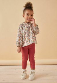 Kind in een witte bloes met bloemenprint, met pofrokken en een gerimpelde halslijn, gecombineerd met geribbelde roze legging en witte sneakers.