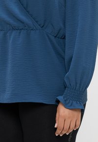 Haut bleu texturé avec une taille froncée, doté de longues manches smockées et d'un ourlet arrondi. Porté avec des leggings noirs.