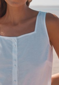 Biely top bez rukávov s hranatým výstrihom a radom gombíkov vpredu. Textília pôsobí ľahko a mäkko s hladkou textúrou.