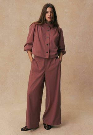Woman with long brown hair wearing a mauve buttoned blouse with puffed sleeves and wide-leg pants, standing with hands in pockets on beige background.