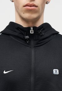 Tuvplāns personai, kas valkā melnu Nike rāvējslēdzēja džemperi ar aukliņām un diviem logotipiem uz krūtīm, redzama apakšējā seja un kakls.