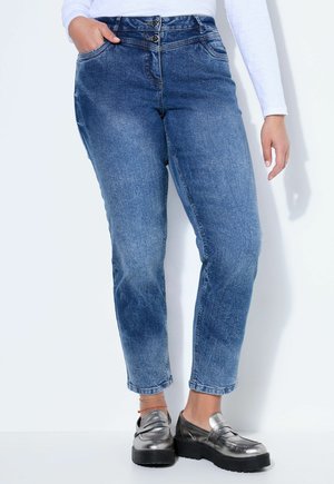 Donna che indossa jeans blu a vita alta con doppi bottoni, top bianco a maniche lunghe e mocassini platform metallici argento, in piedi con una mano in tasca.