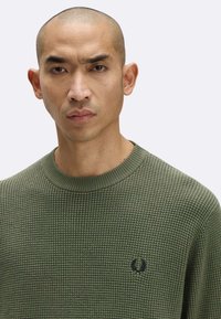 Maglione in maglia waffle verde oliva con collo a giro e polsini a costine; presenta un piccolo emblema scuro sul lato sinistro del petto.