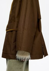 Hnedá bunda z textúrovanej tkaniny, s akcentmi z jemne brúseného materiálu a chlpatým lemom. Voľný strih s viditeľným šitím a detailom na gombíku.