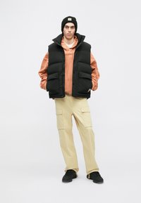 Svart pufferväst över en orange hoodie; beige cargobyxor; svart mössa och sneakers; textil med struktur; flera fickor på västen.
