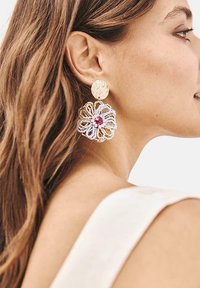 Pendientes circulares en tono dorado con discos texturizados, que presentan detallados diseños de flores de cuentas en blanco y rosa con un acento de gema central.