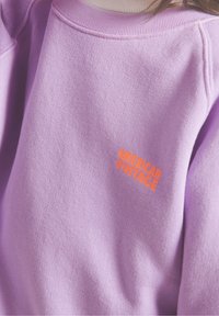 Fioletowa bluza z miękkiego materiału, z prążkowanym dekoltem i pomarańczowym logo "AMERICAN VINTAGE" z przodu.