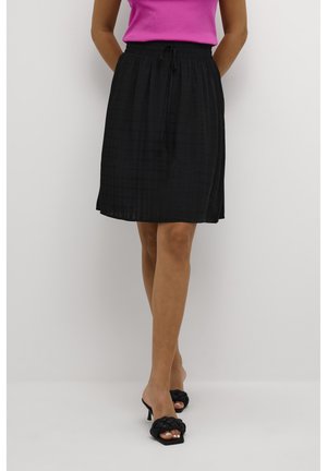 Kobieta ubrana w czarną spódnicę sięgającą kolan z elastycznym pasem oraz różowy, bezrękawnik, zestawione z czarnymi plecionymi sandałami na obcasie.