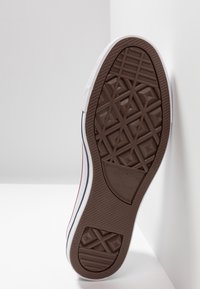 Semelle d'une chaussure classique Converse All Star avec une bande de roulement en caoutchouc marron et une semelle intermédiaire blanche appuyée contre un mur d'angle blanc.