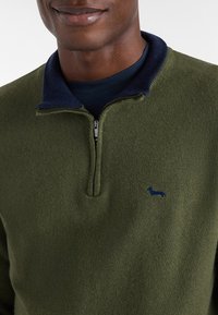 Oliwkowy sweter z granatowym kołnierzem, z ćwierć zamkiem i małym granatowym logo psa na piersi. Miękka, teksturowana tkanina.