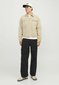 Jachetă din denim bej cu două buzunare pe piept și nasturi tip snap, asociată cu pantaloni cargo negri ce au multiple buzunare și adidași bej.