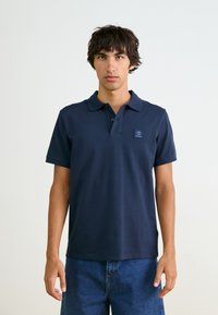 Navy krāsas polo krekliņš no kokvilnas ar apkakli, trīs pogu aizdari un mazu logotipa uzlīmi kreisajā krūtī. Valkāts kopā ar džinsu šortiem.