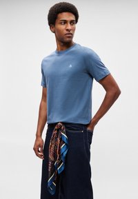 Zils kokvilnas t-krekls ar apaļu kaklu un īsām piedurknēm. Uz krūtīm neliels logotips. Valkāts ar tumšiem džinsa biksēm un rakstainu šalli, kas sasieta apkārt viduklim.