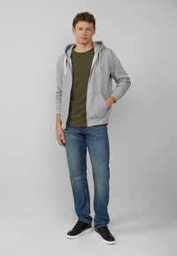 Szary hoodie na zamek z białymi sznurkami, oliwkowa koszulka, niebieskie proste dżinsy i czarne sneakersy z białymi podeszwami.