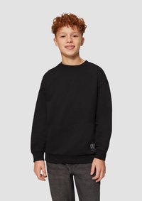 Felpa nera con maniche lunghe e polsini a costine, con un piccolo logo bianco in basso a sinistra. Indossata con jeans scuri.
