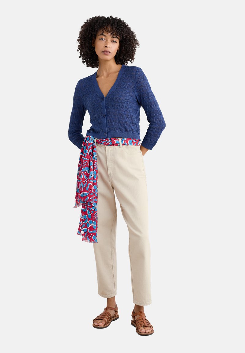 Marinblå stickad cardigan med ett texturerat mönster, beige byxor och en röd och blå blommig scarf knuten i midjan. Bruna sandaler med remmar.