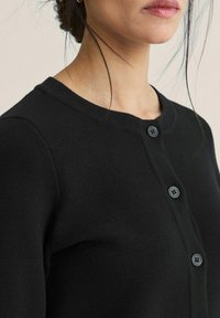 Svart stickad kofta med rundad halsringning, med två framknappar och en texturerad tyg. Nära vy med fokus på knapparna och texturen.