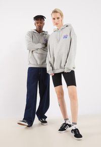 Grå hættetrøjer med frontlommer, båret af to personer. Den ene har mørkeblå løse jeans på; den anden har sorte cykelshorts og sneakers.