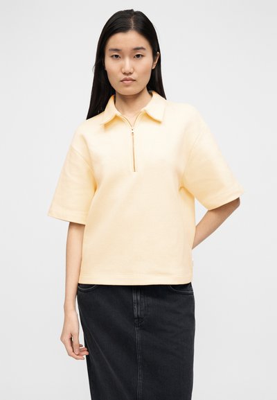 Πόλο με κοντά μανίκια σε ανοιχτό κίτρινο ύφασμα με υφή και λαιμόκοψη με φερμουάρ μέχρι τη μέση, συνδυασμένο με σκουρόχρωμη φούστα από τζιν.