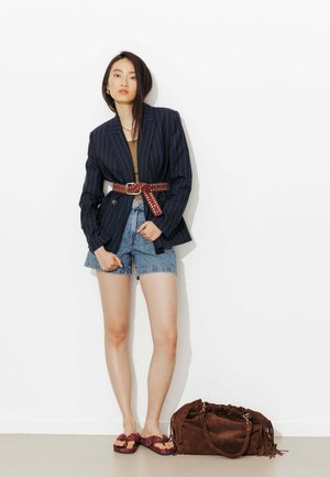 Femme portant un blazer à fines rayures ceinturé d'une ceinture marron cloutée, un short en jean et des sandales, debout à côté d'un grand sac en suède marron à franges.