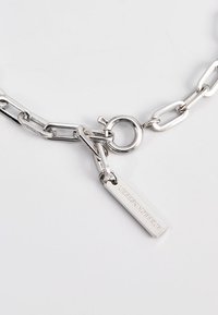 Silberne Kettenhalskette mit verbundenen ovalen und runden Metallstücken, mit einem runden Verschluss und einem rechteckigen Anhänger, der mit Text graviert ist.