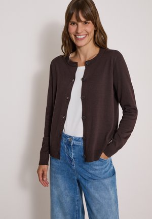 Femme souriante portant un cardigan marron foncé boutonné sur une chemise blanche et un jean bleu, avec une main dans sa poche.