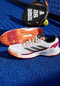 Witte sportschoen met zwarte en grijze accenten, lichtgewicht mesh bovenkant, oranje en rode zool, gestructureerde loopzool en vetersysteem.