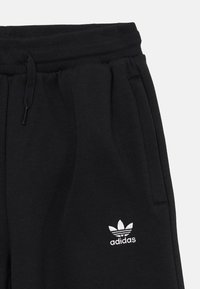 Čierne jogger nohavice vyrobené z mäkkého materiálu, s pásom na šnúrku, bočnými vreckami a bielym vyšívaným logom Adidas na stehne.