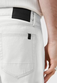 Hvide denim jeans med en sort læderlap mærket "ALLSAINTS" på ryggen. Funktioner inkluderer rene linjer, baglomme og kontrastsy. 