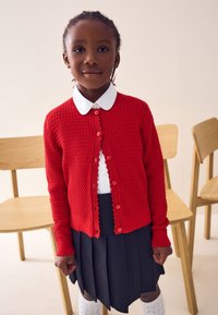 Rdeč pleteni kardigan z teksturiranim vzorcem, valovitim robom in gumbi. Združen z belo srajco z ovratnikom in črno, nagubano krilo.