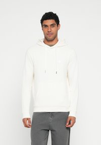 Kremowa bluza z kapturem, przednią kieszenią, sznurkiem przy kapturze i subtelnym logo. Miękki materiał i ściągacze przy rękawach, zestawiona z szarymi spodniami.