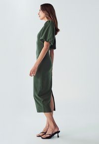 Grünes Midikleid mit Puffärmeln, seitlichem Schlitz und figurschmeichelndem Design. Kombiniert mit schwarzen Absatzsandalen, vor einem neutralen Hintergrund.